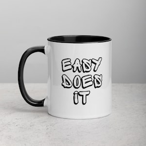 Puede incluir: Taza de café de cerámica blanca con borde y asa negros. La taza tiene el texto "EASY DOES IT" impreso en negro en la parte delantera.