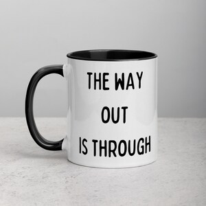 Op de afbeelding: Witte keramische koffiemok met een zwarte rand en handvat. De mok heeft de tekst "The way out is through" in het zwart gedrukt.