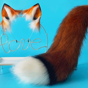 Puede incluir: Un accesorio de disfraz de cola de zorro de peluche con una punta blanca, una secci&oacute;n media negra y una larga secci&oacute;n de piel marr&oacute;n rojiza. La cola est&aacute; unida a una base de madera y se exhibe en un soporte blanco con un letrero de "love" de metal dorado.