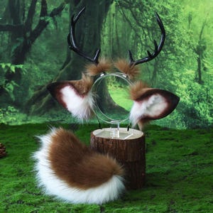 May include: A deer-themed costume set featuring a headband with antlers and ears, and a fluffy tail. The antlers are black, and the ears are brown with white and brown accents. The tail is brown with a white tip. The set is displayed on a wooden stand against a forest backdrop.