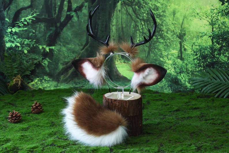 May include: A deer-themed costume set featuring a headband with antlers and ears, and a fluffy tail. The antlers are black, and the ears are brown with white and brown accents. The tail is brown with a white tip. The set is displayed on a wooden stand against a forest backdrop.