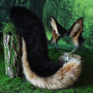 May include: A black, tan, and cream-colored faux fur fox tail and matching ear headband set. The tail is long and fluffy, with a black top and a tan and cream-colored bottom. The ears are black with tan and cream-colored fur.