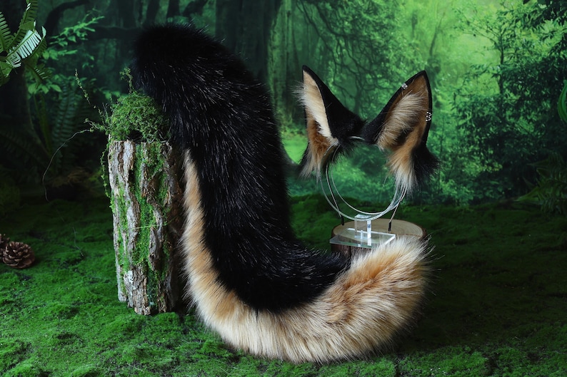 May include: A black, tan, and cream-colored faux fur fox tail and matching ear headband set. The tail is long and fluffy, with a black top and a tan and cream-colored bottom. The ears are black with tan and cream-colored fur.