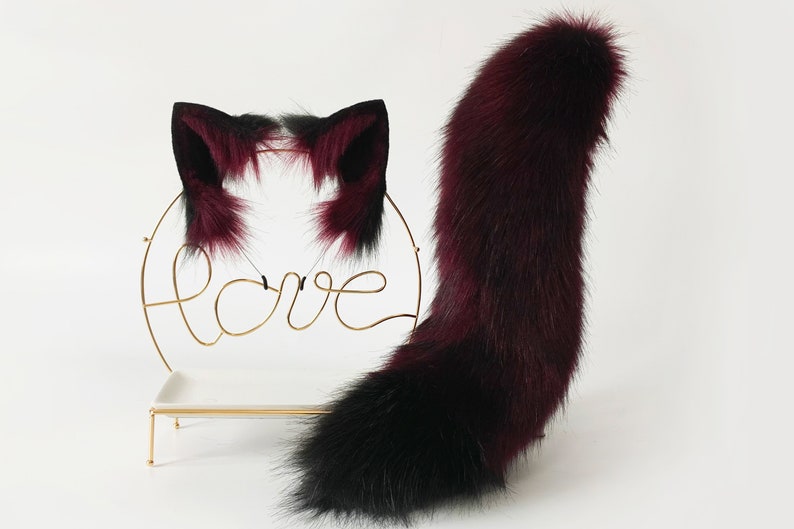 May include: A set of burgundy and black faux fur cat ears and tail. The ears are attached to a headband and the tail is long and fluffy.