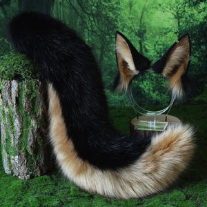 May include: A black, tan, and cream-colored faux fur fox tail and matching ear headband set. The tail is long and fluffy, and the ears are detailed with tan and black accents. The set is displayed on a green mossy surface.