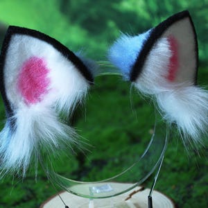 May include: A pair of cat ear headbands. Each ear is white with a pink inner ear and a black trim. The ears have a fluffy white fur accent and are attached to a clear plastic headband.