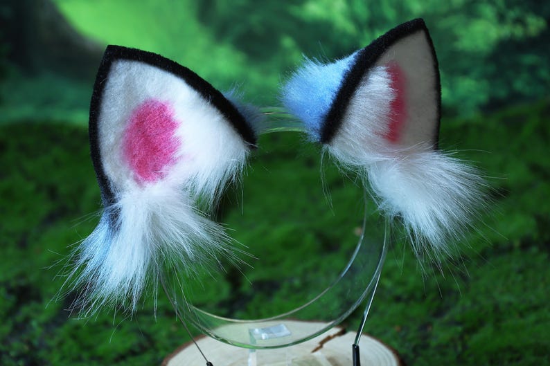 May include: A pair of cat ear headbands. Each ear is white with a pink inner ear and a black trim. The ears have a fluffy white fur accent and are attached to a clear plastic headband.