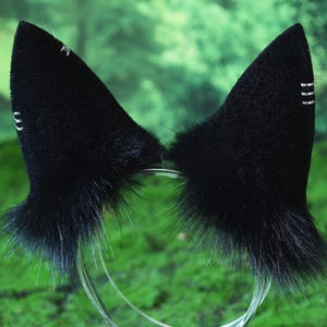 May include: Black cat ear headband with fluffy fur and silver accents. The ears are mounted on a clear plastic headband. The ears have a pointed shape and are set against a blurred green background.