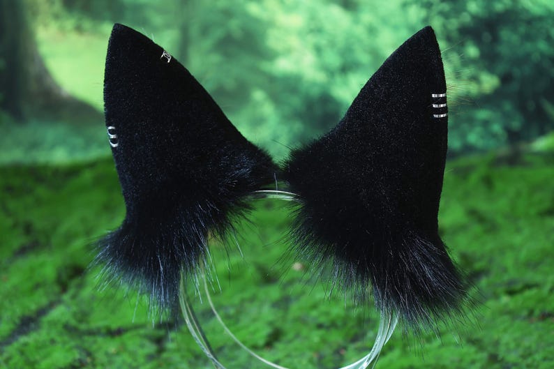 May include: Black cat ear headband with fluffy fur and silver accents. The ears are mounted on a clear plastic headband. The ears have a pointed shape and are set against a blurred green background.