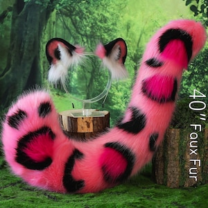 May include: A pink faux fur tail, approximately 40 inches long, with black spots. A matching headband with ears is also included. The tail is displayed on a green surface with a forest backdrop.