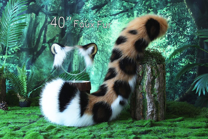 May include: A 40-inch long faux fur tail with a black, white, and brown spotted pattern. The tail is curled up on a mossy green surface with a blurred background of green foliage.