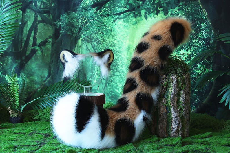 May include: A fluffy, brown, black, and white cat tail with a spotted pattern. The tail is curled up and appears to be made of faux fur. The tail is in front of a green and brown background.