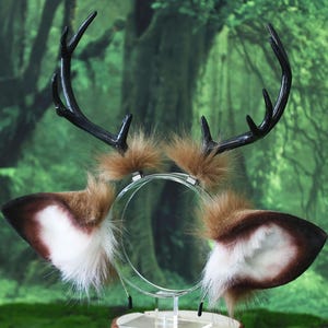 May include: A whimsical deer antler and ear headband. The antlers are black, and the ears are brown and white with fluffy accents. The headband is clear, and the background is a blurred green forest.