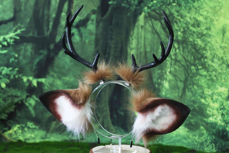 May include: A whimsical deer antler and ear headband. The antlers are black, and the ears are brown and white with fluffy accents. The headband is clear, and the background is a blurred green forest.