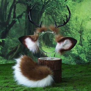May include: A deer-themed costume set featuring a headband with antlers, ear attachments, and a tail. The antlers are black, and the ears are brown with white and brown fur. The tail is brown and white. The set is displayed on a wooden stand against a green background.