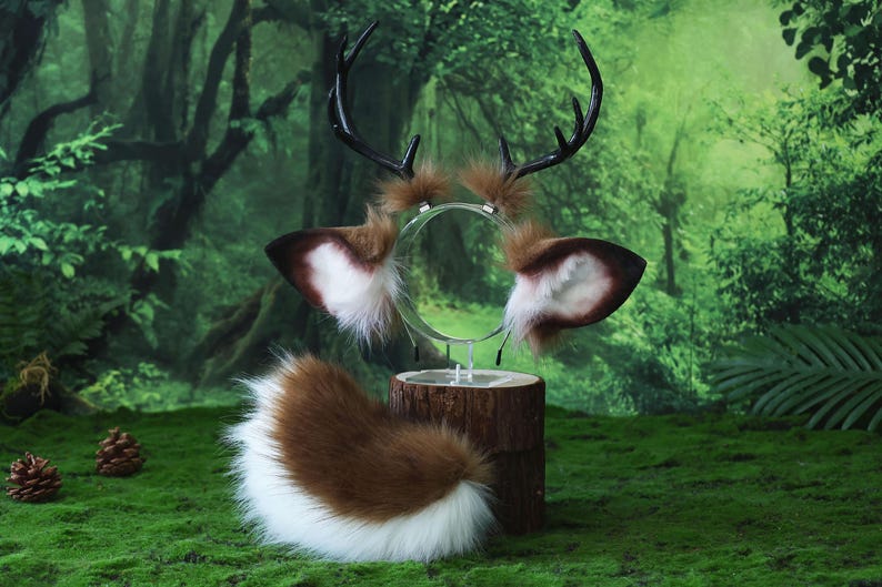 May include: A deer-themed costume set featuring a headband with antlers, ear attachments, and a tail. The antlers are black, and the ears are brown with white and brown fur. The tail is brown and white. The set is displayed on a wooden stand against a green background.