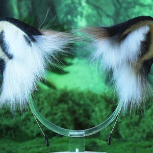 May include: A pair of white and black faux fur cat ears with a clear plastic headband. The ears are attached to the headband with black wire.