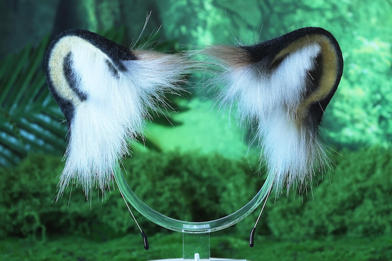 May include: A pair of white and black faux fur cat ears with a clear plastic headband. The ears are attached to the headband with black wire.
