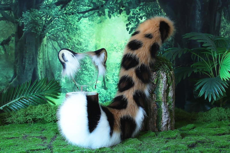May include: A brown, black, and white faux fur tail with a matching headband featuring cat ears. The tail is curled up and the headband is on a stand. The background is a green forest.