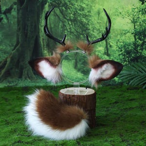 May include: A deer-themed costume set featuring a headband with antlers and ears, and a fluffy tail. The antlers are black, and the ears are brown and white. The tail is brown and white. The set is displayed on a wooden stand against a green background.