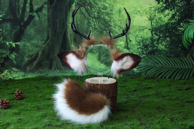 May include: A deer-themed costume set featuring a headband with antlers and ears, and a fluffy tail. The antlers are black, and the ears are brown and white. The tail is brown and white. The set is displayed on a wooden stand against a green background.
