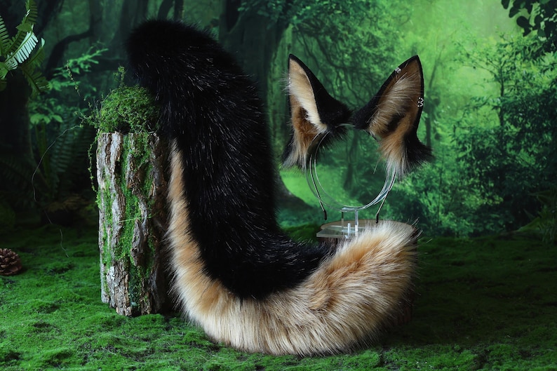 May include: A black, tan, and white fox tail and ear set. The tail is long and fluffy, with a gradient of colors. The ears are attached to a clear headband. The set is displayed on a bed of green moss, with a forest background.