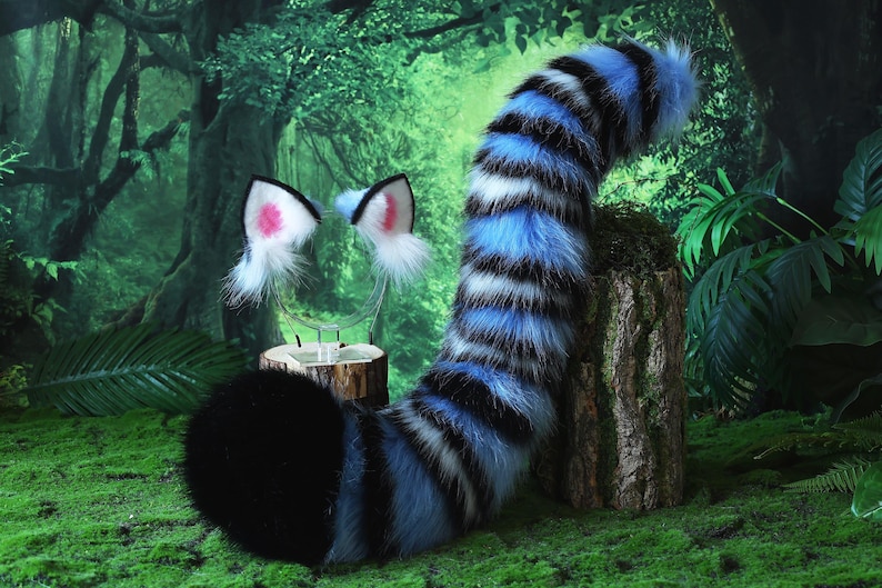 May include: A furry costume set featuring a black and blue striped tail and matching cat ears with pink inner ears. The tail is propped up on a wooden stump against a backdrop of a lush green forest.