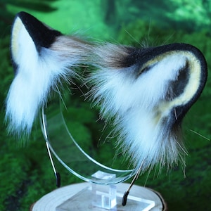 May include: A pair of faux fur cat ears headband in black, white, and brown with a yellow accent. The headband is on a clear stand.