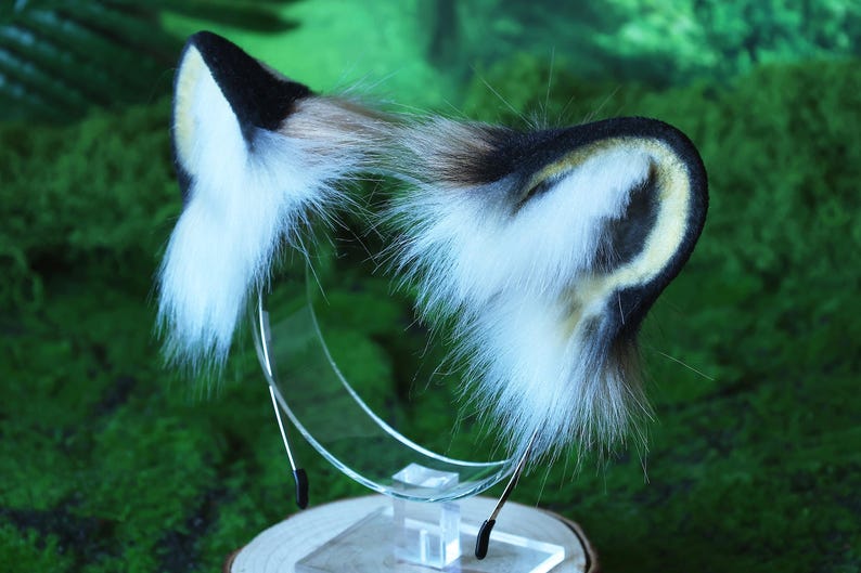 May include: A pair of faux fur cat ears headband in black, white, and brown with a yellow accent. The headband is on a clear stand.