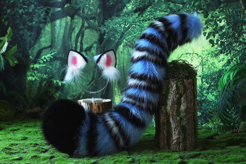 May include: A furry costume tail and cat ear headband set. The tail is blue, black, and white striped, and the ears are white with pink inner ears. The items are displayed on a mossy surface with a forest backdrop.