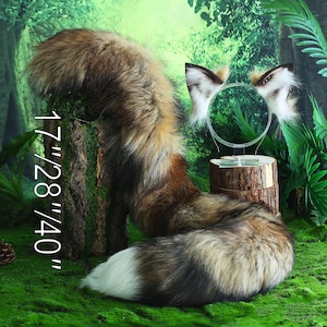 Puede incluir: Una cola de zorro esponjosa de color marrón y blanco con una punta blanca, que mide 17/28/40 pulgadas. La cola se muestra en un tocón de árbol en un entorno de bosque con follaje verde.