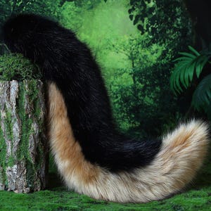 May include: A faux fur tail in black and tan, resting on a green surface. The tail is thick and fluffy, with a gradient of colors. The background features a green forest scene.