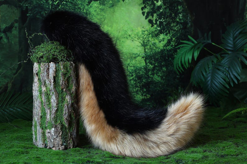 May include: A faux fur tail in black and tan, resting on a green surface. The tail is thick and fluffy, with a gradient of colors. The background features a green forest scene.