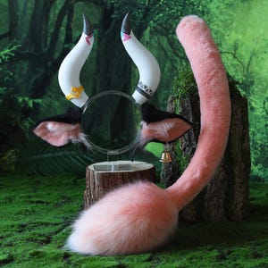 May include: A whimsical costume set featuring a headband with white horns, black ears with pink insides, and a fluffy pink tail. The horns have black tips and decorative accents. A small gold bell hangs from one ear. The set is displayed on a mossy surface.