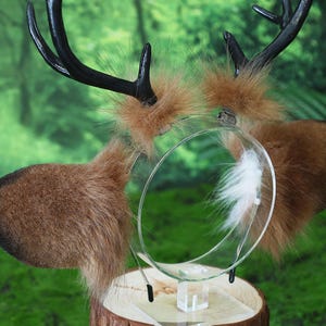 May include: A deer antler headband with brown faux fur ears and black antlers. The headband has a clear plastic band with a white feather accent. The ears are a light brown color and the antlers are black.
