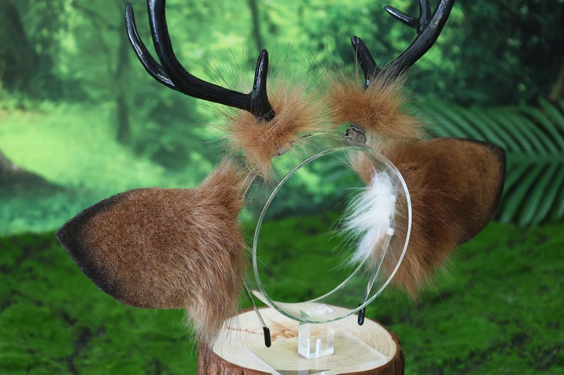 May include: A deer antler headband with brown faux fur ears and black antlers. The headband has a clear plastic band with a white feather accent. The ears are a light brown color and the antlers are black.