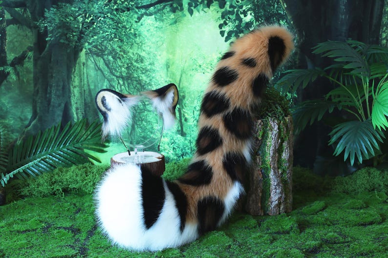 May include: A brown, black, and white faux fur tail with a spotted pattern. The tail is curled up and is sitting on a green mossy surface.