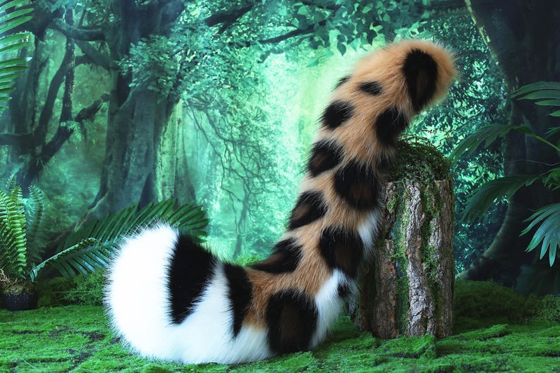 May include: A long, fluffy, brown, black, and white faux fur tail with a spotted pattern. The tail is curled up on a mossy green surface.
