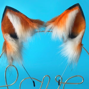 Puede incluir: Un par de orejas de zorro de piel sint&eacute;tica naranja y blanca con detalles negros. Las orejas est&aacute;n unidas a una diadema de metal dorado.