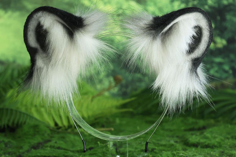 May include: A pair of white and black faux fur animal ears with a black headband. The ears are attached to a clear plastic headband with black metal clips.