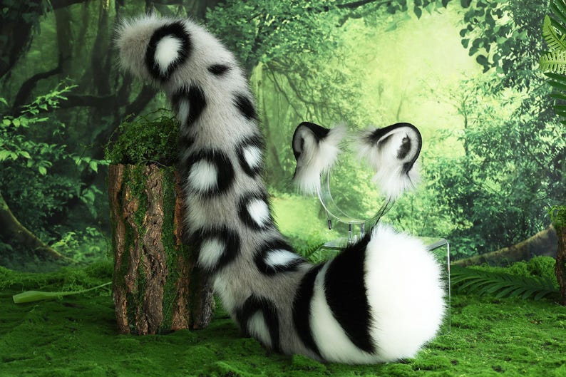 May include: A white and black spotted faux fur tail with a white tip. The tail is curled around a white faux fur headband with black ears. The tail and headband are on a green mossy surface.