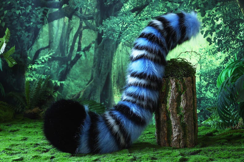 May include: A plush, blue, black, and white striped tail prop, likely for cosplay or costume use, rests on a moss-covered tree stump. The tail is set against a backdrop of a lush, green forest, creating a whimsical scene.