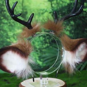May include: A deer antler headband with brown and white faux fur ears and black antlers. The ears are attached to a clear plastic headband. The antlers are black and curved. The headband is displayed on a wooden stand.