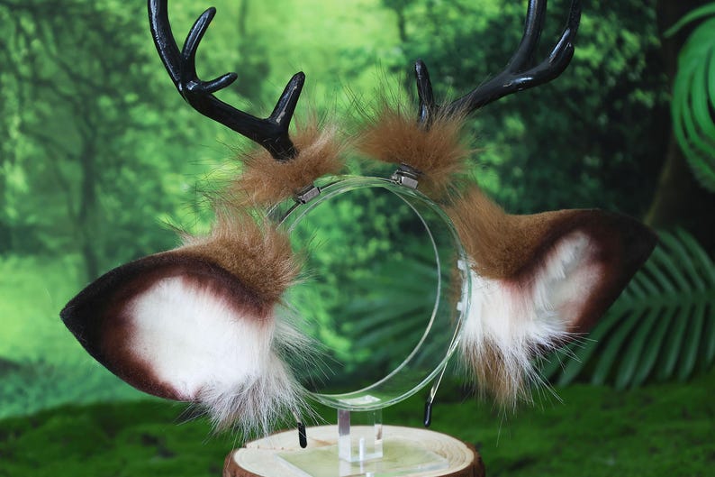 May include: A deer antler headband with brown and white faux fur ears and black antlers. The ears are attached to a clear plastic headband. The antlers are black and curved. The headband is displayed on a wooden stand.