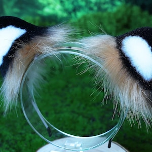 May include: A black and white furry headband with a brown fur trim. The headband is made of soft, plush fabric and is designed to look like animal ears.