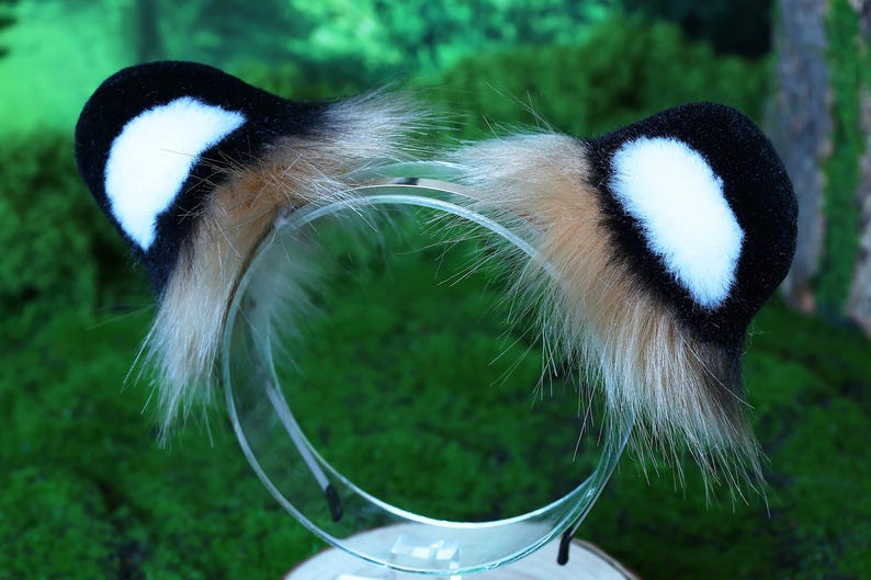 May include: A black and white furry headband with a brown fur trim. The headband is made of soft, plush fabric and is designed to look like animal ears.