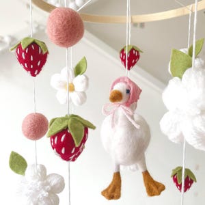 May include: A nursery mobile with felt strawberries, pink wool balls, white flowers, and a goose wearing a pink bonnet. The mobile is suspended from a wooden hoop with white string.