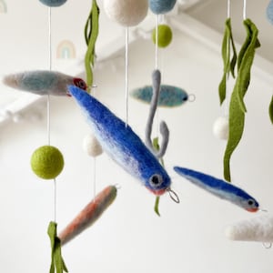 Op de afbeelding: Handgemaakte baby mobile met vilten vissen in blauw, grijs en oranje, samen met groen zeewier en witte en blauwe bollen. De mobile hangt aan een houten ring, een grillige decoratie voor de kinderkamer.