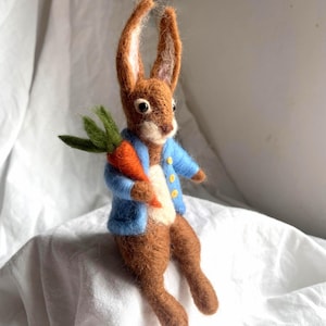 Puede incluir: Una figura de conejo de fieltro hecha a mano con orejas largas y erguidas. El conejo lleva una chaqueta azul claro con botones amarillos y sostiene una zanahoria naranja con hojas verdes. El conejo es marrón con el pecho color crema.
