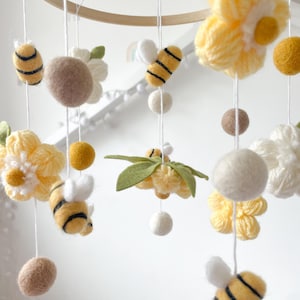 May include: A hanging mobile featuring yellow and black felt bees, white and yellow yarn flowers, and solid felt balls in shades of white, tan, and mustard yellow. The elements are suspended by white strings from a circular wooden frame.